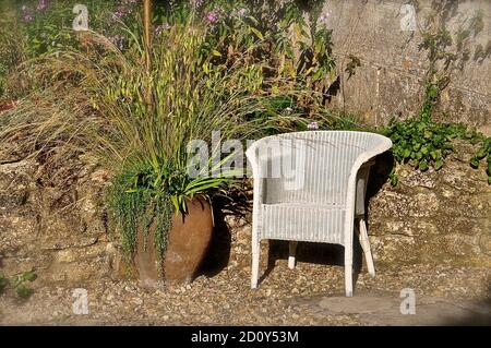 Versunkene Terrasse aus Steinsplitter und einer Stützsteinmauer in einem englischen Landgarten, mit einem weißen Lloyd Loom Korbstuhl neben einer großen Terrasse Stockfoto