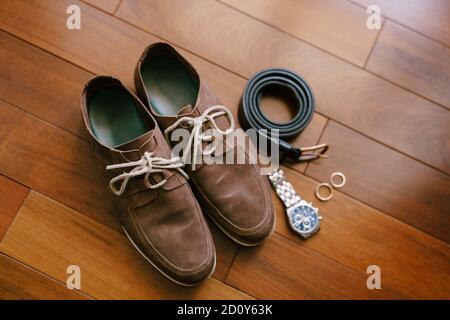 Holzboden mit braunen Herrenschuhen, Armbanduhr, Gürtel und Eheringen. Stockfoto