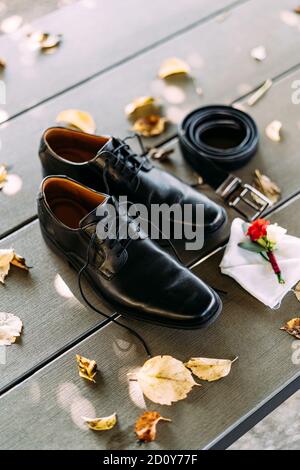 Schwarze Schuhe eines Mannes mit unbanden Schnürsenkeln auf einer Holzstruktur mit einem Gürtel und Boutonniere des Bräutigams auf einem weißen Taschenquadrat. Stockfoto