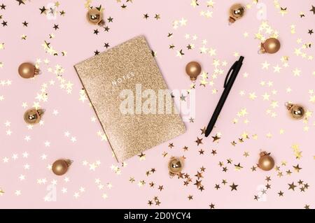 Goldenes Notizbuch mit glänzendem Glitter-Cover, schwarzem Stift und goldenen Weihnachtskugeln, Kugeln und Konfetti-Sternen auf rosa Hintergrund. Wunschliste, Neujahrsreso Stockfoto