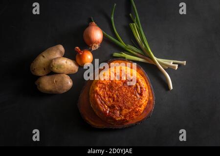 Blick von oben auf ein Kartoffelomelett auf einem Holzbrett Neben Kartoffeln und Zwiebeln auf schwarzem Hintergrund Stockfoto