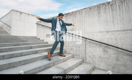 Fröhlicher und glücklicher Geschäftsmann in einem Anzug ist Kaffee halten und aktiv tanzen, während Sie die Treppe hinunter gehen. Szene aufgenommen in einem städtischen Betonpark Stockfoto