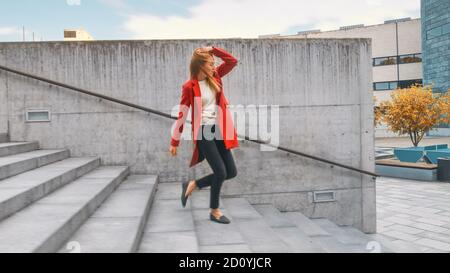 Fröhliche und glückliche junge Frau aktiv tanzen beim Gehen die Treppe hinunter. Sie trägt einen langen roten Mantel. Szene aufgenommen in einem städtischen Betonpark Stockfoto