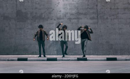 Multiple Exposure Shot eines coolen geklonten jungen Hipstermannes mit langen Haaren steht in Tanzposen auf einer Straße neben einer großen Betonwand. Er trägt einen Stockfoto