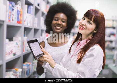 Erfahrene professionelle Apothekerin hält Tablette und berät afrikanische Kundin in der modernen Apotheke. Apotheker und Patient bei Modern Stockfoto