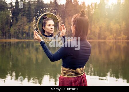 Herbst-Bohemian Foto einer Frau mit langen Haaren hält einen runden goldenen Spiegel mit Spiegelung ihres Gesichts. Stockfoto