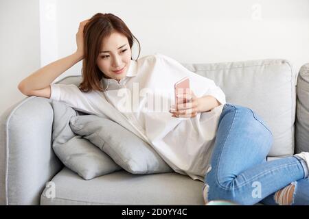 Junge asiatische Frau liegend entspannend auf Couch Blick auf mobile Telefon Stockfoto