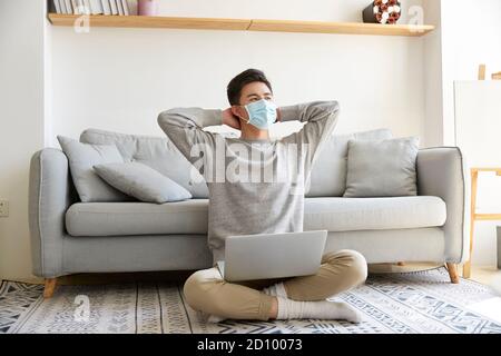 Junge asiatische Geschäftsmann zu Hause mit Maske sitzen bleiben Auf dem Teppich, der mit einem Laptop gearbeitet hat Stockfoto