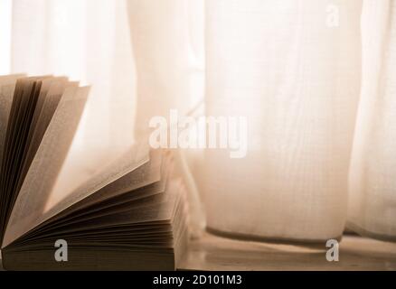 Offenes Buch auf der Fensterbank, mit Vorhängen und warmem Licht Stockfoto