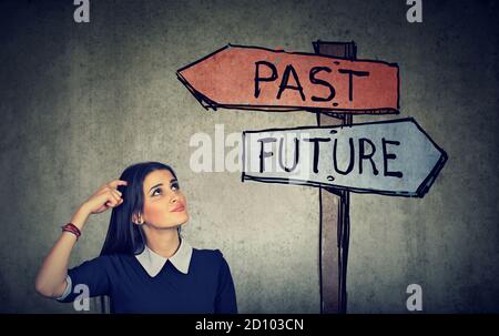 Nachdenkliche junge Frau, die auf einen Wegweiser mit Pfeilen schaut Zeigt vergangene und zukünftige Richtungen an Stockfoto