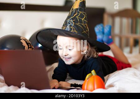 Lustige Mädchen im Hexenkostüm für Halloween liegt auf dem Bett und mit einem digitalen Tablet Laptop Notebook. Online-Anruf bei Freunden oder Eltern. Stockfoto