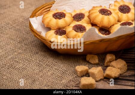 Hausgemachte Gebäck-Kekse mit Marmelade in einem Weidenkorb und Stücke von braunem Rohrzucker auf einem Hintergrund von homespun Stoff mit einer rauen Textur, close-up, Stockfoto