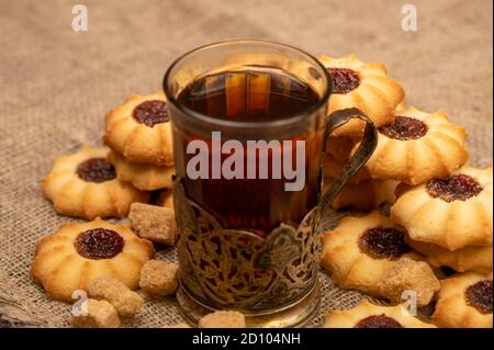 Hausgemachte Gebäck-Kekse mit Marmelade und einem facettierten Glas Tee in einem Vintage-Cup-Halter auf einem Hintergrund von homespun Stoff mit einer rauen Textur, close-up, Stockfoto