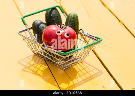 Lustige rote Tomate mit Augen und grünen Gurken im kleinen Supermarkt Korb auf dem gelben Hintergrund. Vegetarisches Konzept, Kopierraum Stockfoto