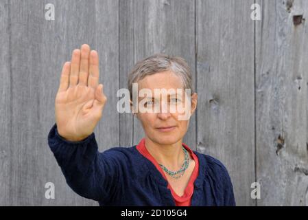 Wütende kaukasische Landfrau mittleren Alters mit kurzen Haaren macht das Handpalmenstoppschild mit ihrer rechten Hand, vor einem alten Scheunenholzhintergrund. Stockfoto