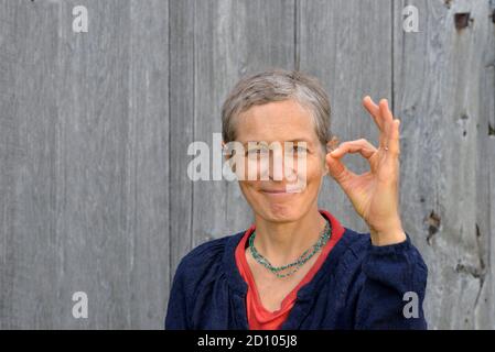 Optimistische, moderne kaukasische Landfrau mittleren Alters zeigt mit der linken Hand das OK-Zeichen (Zustimmungsgeste) vor altem Scheunenholzhintergrund. Stockfoto