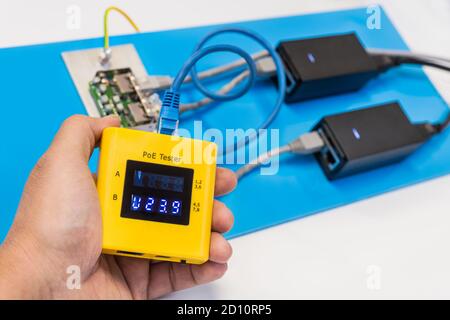 Messung der PoE-Netzspannung durch Datenkabeltester in der Hand eines Technikers. Zwei geschaltete Netzteile, elektrische Drähte und ein Überspannungsschutz auf blauem Kunststoff. Stockfoto