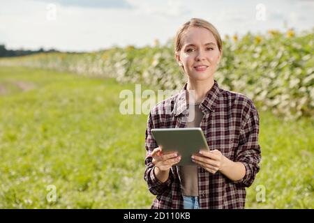 Junge blonde lächelnde Farmerin in Arbeitskleidung mit Tablet gegen Grünes Feld Stockfoto