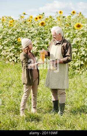 Happy Senior männlichen Landwirt in Arbeitskleidung vorbei Flasche Sonnenblume Öl zu weiblich Stockfoto