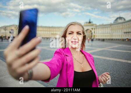 Russische Frau 37 Jahre alt nimmt ein Selfie in St. Petersburg, Russland. Lächelnde Touristin in rosa fotografiert sich mit einem Smartphone. Stockfoto