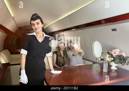 Air Hostess bedient Passagiere im Flugzeug. Schöne Flugbegleiterin in der Passagierkabine des Flugzeugs auf dem Hintergrund von jungen Frauen in Stockfoto