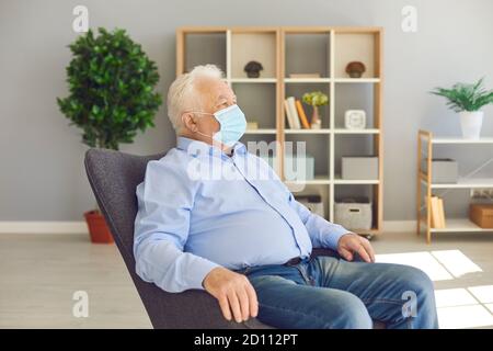 Der ältere Mann sitzt in einer schützenden medizinischen Maske auf seinem Gesicht in einem hellen Raum. Stockfoto