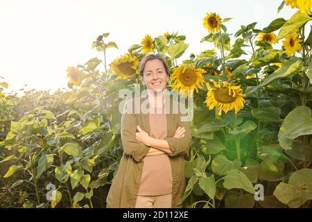 Glücklich reife Frau in Casualwear überqueren ihre Arme auf der Brust Unter Sonnenblumen Stockfoto