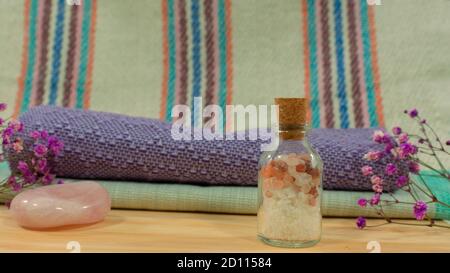 Zusammensetzung mit Glasflasche mit Badesalz auf Holztisch mit Handtüchern, rosa Stein, lila Blumen und Tuch mit Linien im Hintergrund Stockfoto