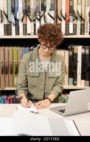 Junge, moderne Verkäuferin, die Notizen im Notizbuch vor sich macht Des Laptops Stockfoto