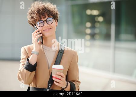 Glückliche junge elegante Geschäftsfrau mit Getränk hält Smartphone am Ohr Stockfoto