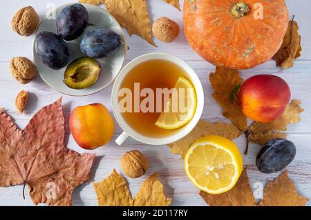 Herbsthintergrund von trockenen Blättern mit einer Tasse Tee, einem kleinen schönen Kürbis, Zitrone, reifen Pflaumen und Nektarinen, Walnüssen. Stockfoto