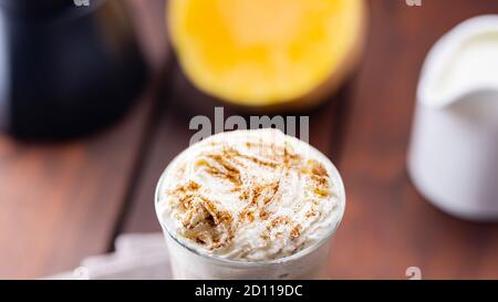 Kürbislatte, Milchkännchen und ein Stück Kürbis. Kürbislatte mit Schlagsahne und Gewürzen auf Holzbrettern. Heißer Herbstkaffee auf einem Laken-Nickerchen Stockfoto
