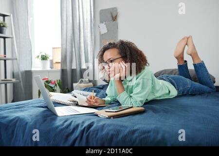 Junge clevere Mixed-Race-Schüler beobachten Online-Unterricht beim Notizen machen Stockfoto
