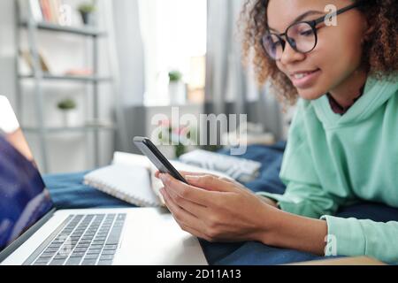 Junge Mischrasse Weibchen in Brillen scrollend in Smartphone während liegend Auf dem Bett Stockfoto