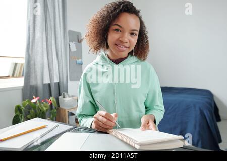 Fröhlicher Teenager-Student mit gemischter Rasse, der im Schlafzimmer Notizen im Copybook macht Stockfoto