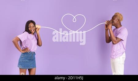 Afroamerikanischer Kerl teilt Gefühle mit seiner Freundin durch Zinn Can-Telefon auf lila Hintergrund Stockfoto