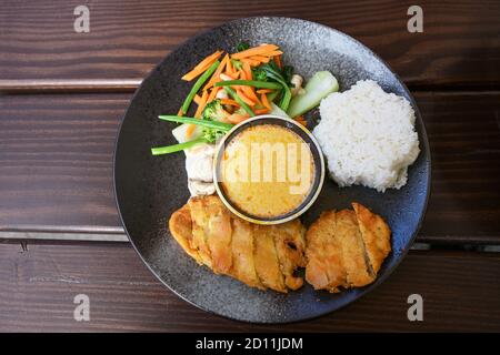 Asiatisches Gericht mit gebratenem Huhn, Gemüse, gedämpftem Reis und Kokosnuss-Curry-Sauce auf dunkelgrauem Teller mit Essstäbchen auf einem Holztisch, Blick aus der Höhe Stockfoto