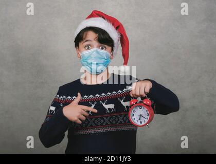 Frohe Weihnachten, Lustiges Kind mit medizinischer Maske und Wecker trägt Weihnachtsmann Hut auf grauem Hintergrund Stockfoto