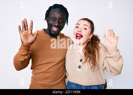 Interracial paar in legerer Kleidung waiving sagen Hallo glücklich und lächelnd, freundliche Willkommensgeste Stockfoto