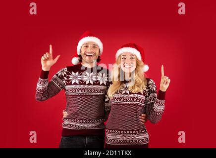 Fröhliche Dame und ihr Freund in Weihnachtspullover und Weihnachtsmann Hüte zeigen auf roten Hintergrund Stockfoto