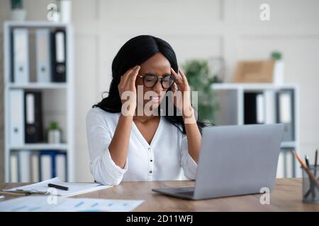 Müde tausendjährige schwarze Geschäftsfrau leiden unter schrecklichen Kopfschmerzen am Schreibtisch Im Büro Stockfoto
