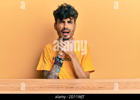 Junge hispanische Mann trägt T-Shirt mit Glück Wort Nachricht auf dem Tisch sitzen schreien und ersticken, weil schmerzhafte Erdrosseln. Gesundheitsproblem. As Stockfoto