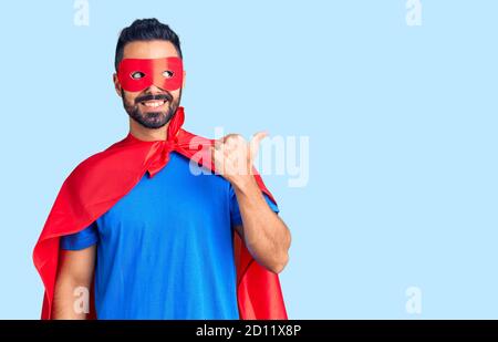Junger hispanischer Mann trägt Superhelden Kostüm lächelnd mit glücklichem Gesicht suchen und zeigen zur Seite mit Daumen nach oben. Stockfoto