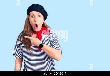 Junge schöne blonde Frau trägt französisch Baskenmütze und gestreiften T-Shirt überrascht mit Finger zur Seite, offenen Mund erstaunt Ausdruck. Stockfoto