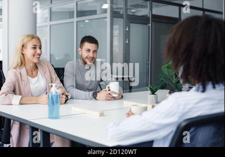 Teamarbeit während der Kaffeepause auf der Toilette im Büro. Europäischer Kerl und Frau mit Tassen, im Gespräch mit afroamerikanischen Dame durch Schutzglas Stockfoto