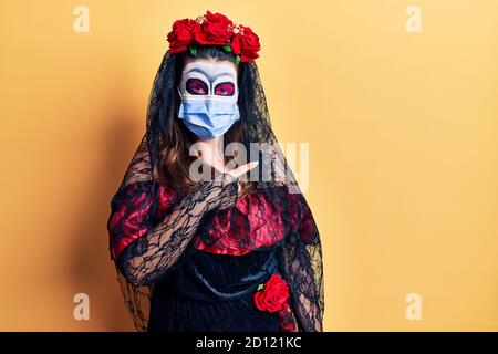 Junge Frau trägt Tag der Toten Kostüm trägt medizinische Maske lächelnd glücklich zeigt mit Hand und Finger auf die Seite Stockfoto