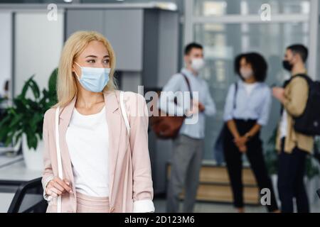 Nach Sperrung und neuer Normalität wieder zur Arbeit zurückkehren. Frau in Schutzmaske sieht zur Seite im Korridor des modernen Büros mit verschwommenen Kollegen Stockfoto