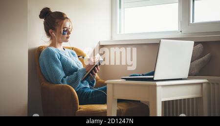 Kaukasische Frau mit roten Haaren und Sommersprossen schreibt etwas In einem Buch mit einem Laptop und Hydrogel Augenflecken Stockfoto