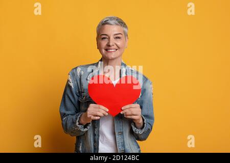 Lächelnd reife Frau hält rotes Papier Herz in der Nähe der Brust Stockfoto