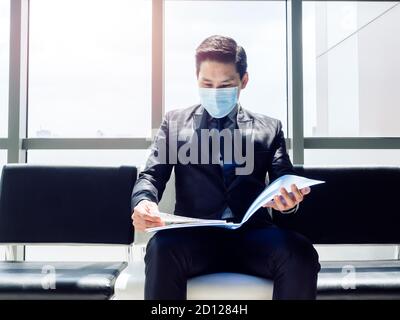 Asiatische Geschäftsmann in Anzug tragen schützende Gesichtsmaske sitzen und Überprüfen Sie seinen Lebenslauf und wartet auf Vorstellungsgespräch in der Nähe riesig Glasfenster im modernen BU Stockfoto